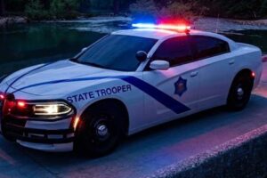 A white State Trooper Dodge Charger police car with its red and blue emergency lights flashing is parked on a concrete structure next to a river at night. The car faces the viewer, and a bridge is visible in the dark background with trees.