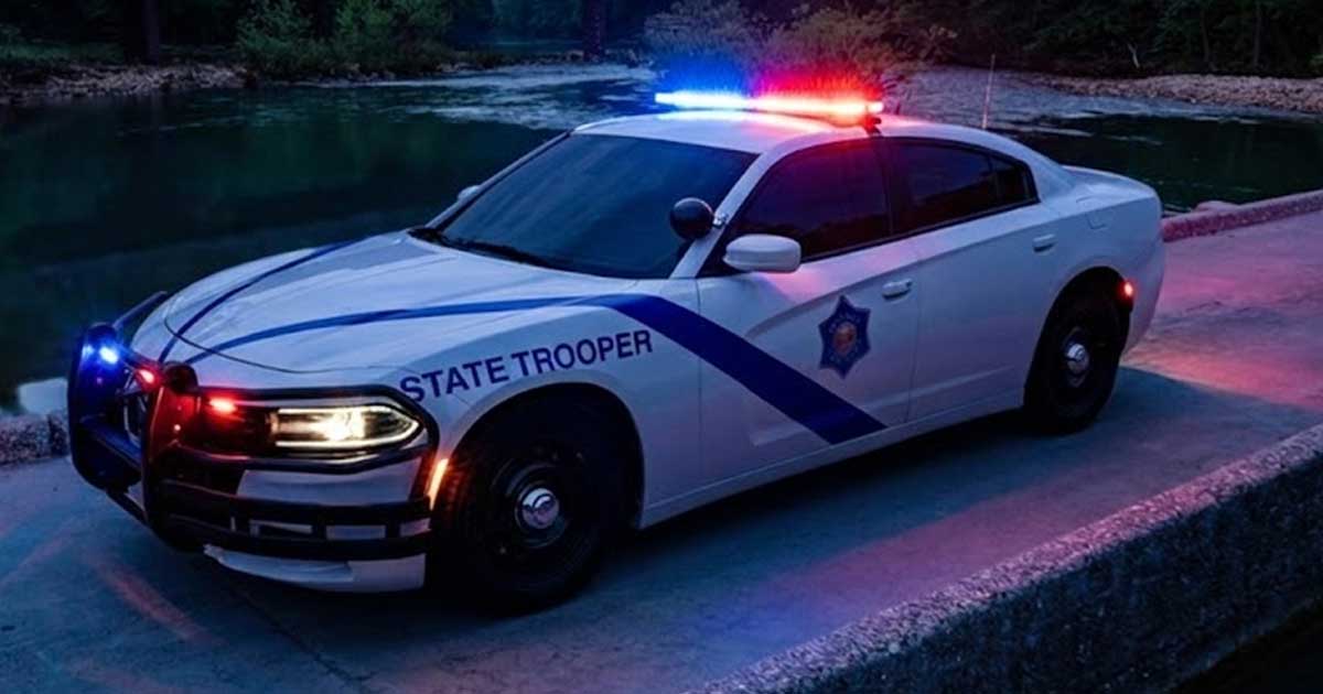 A white State Trooper Dodge Charger police car with its red and blue emergency lights flashing is parked on a concrete structure next to a river at night. The car faces the viewer, and a bridge is visible in the dark background with trees.